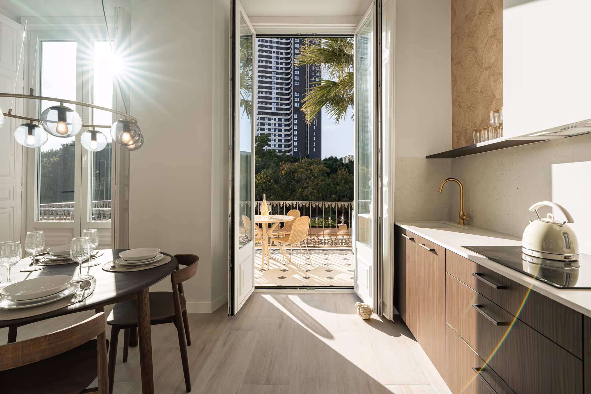 Sunlight streams into the dining area and kitchen of the Aurum residence, with dark wood furniture and light wood floors. Open doors lead to a furnished balcony with city views