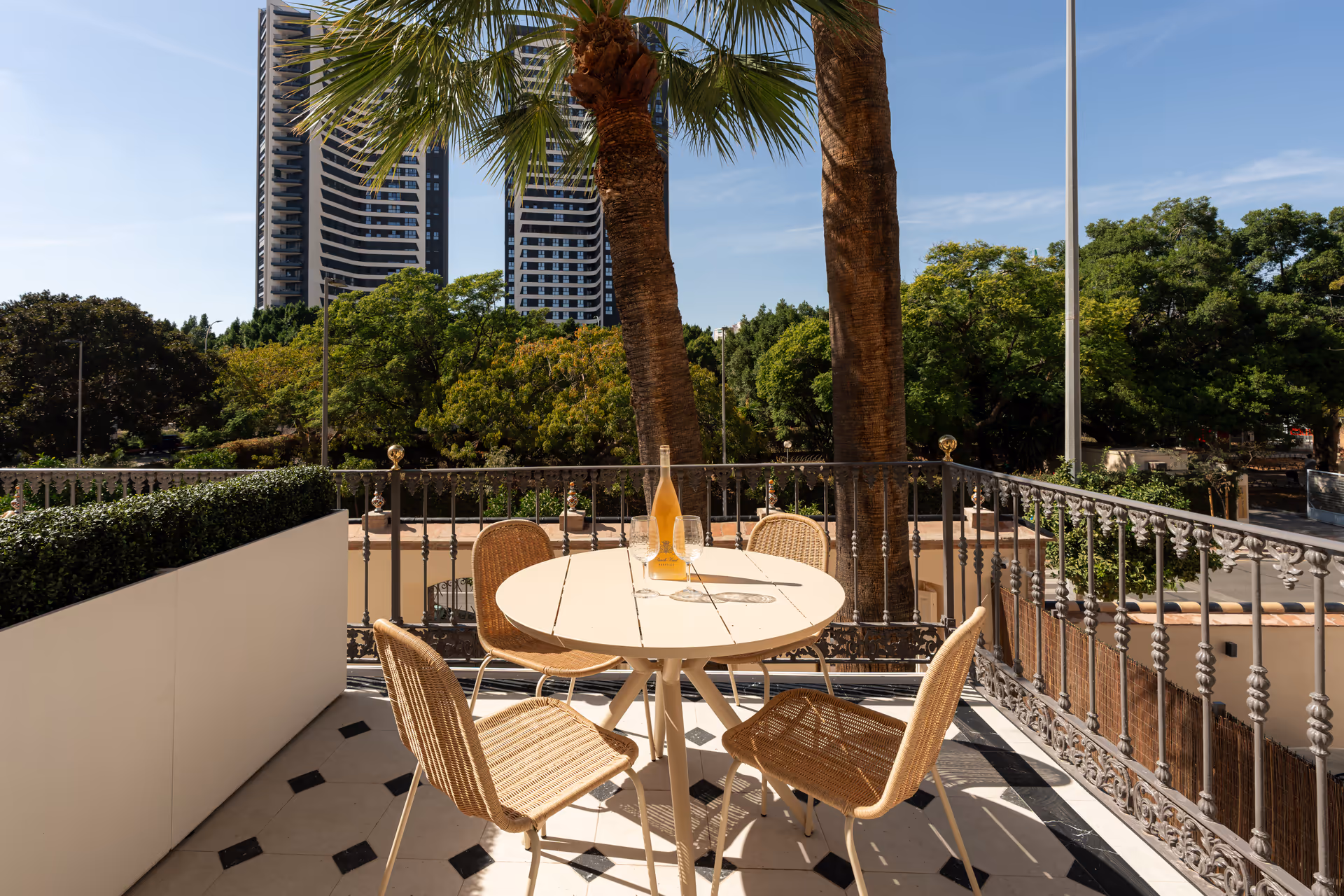 The balcony of the Azuri residence has a round table and four wicker chairs, overlooking palm trees and modern Málaga towers