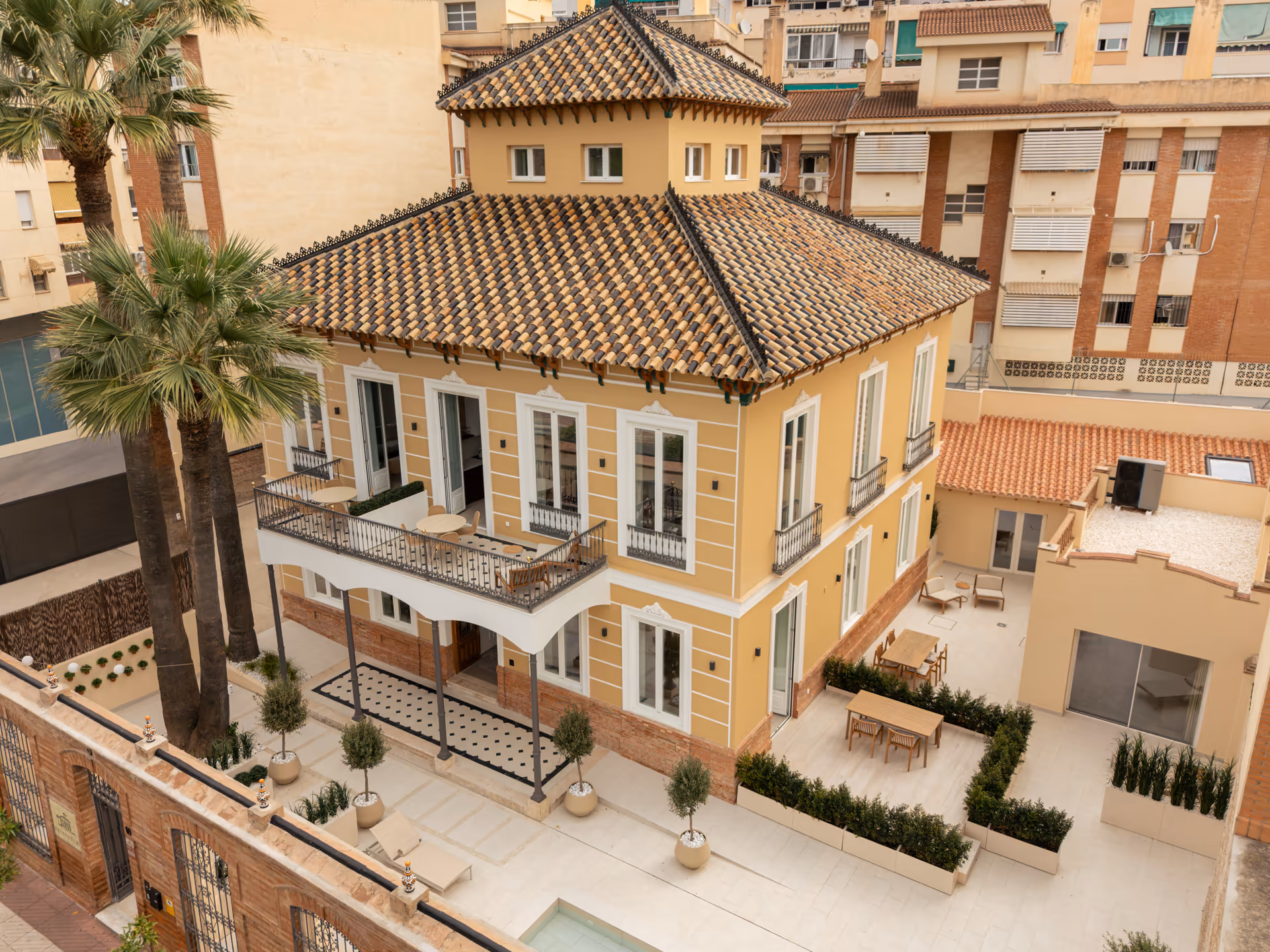 An aerial view of Palacete 10, a restored yellow villa with a tiled roof, tower, and terraces, surrounded by taller buildings in Málaga