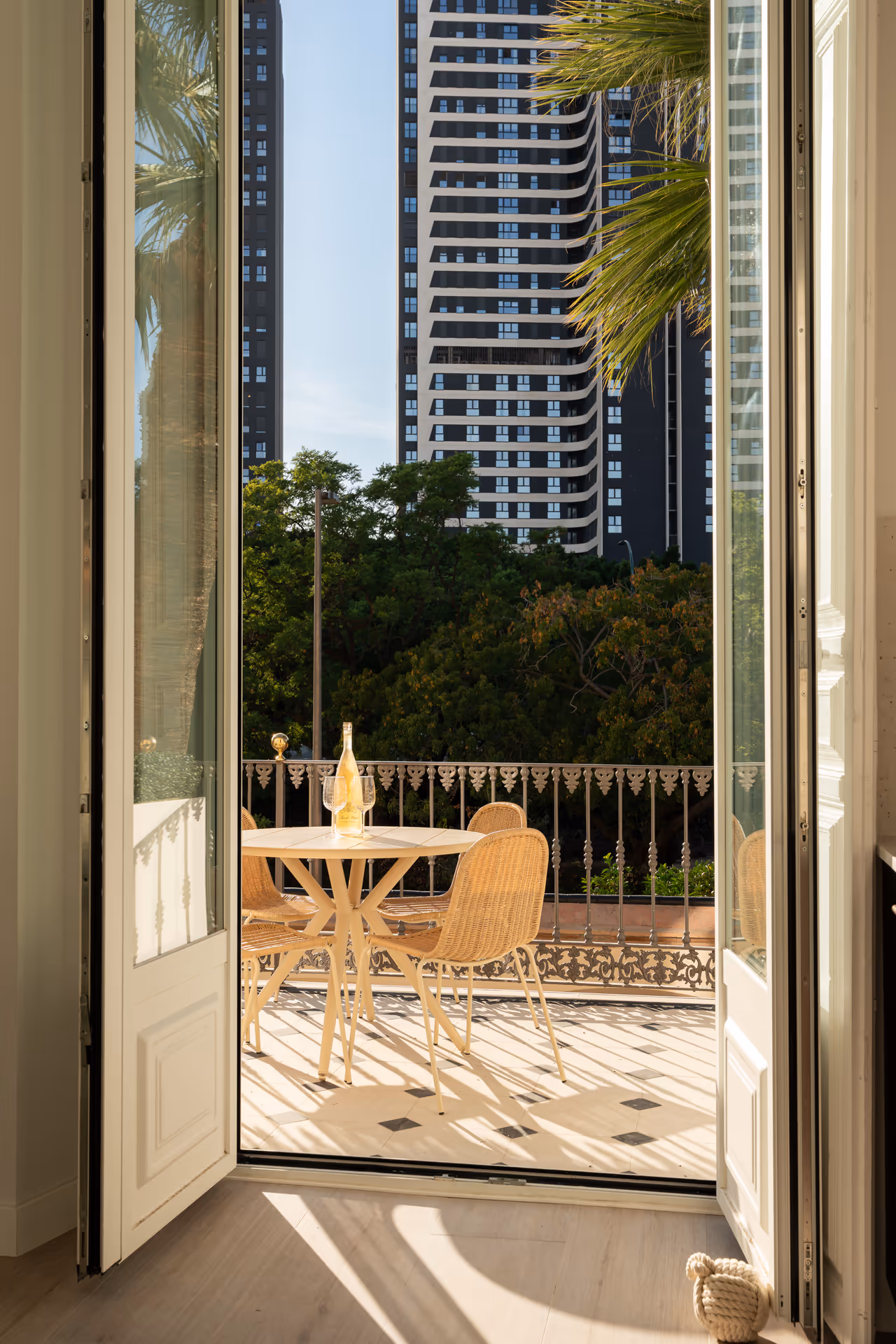 Open doors from the Aurum residence lead to a sunny terrace with a dining set, overlooking trees and modern buildings