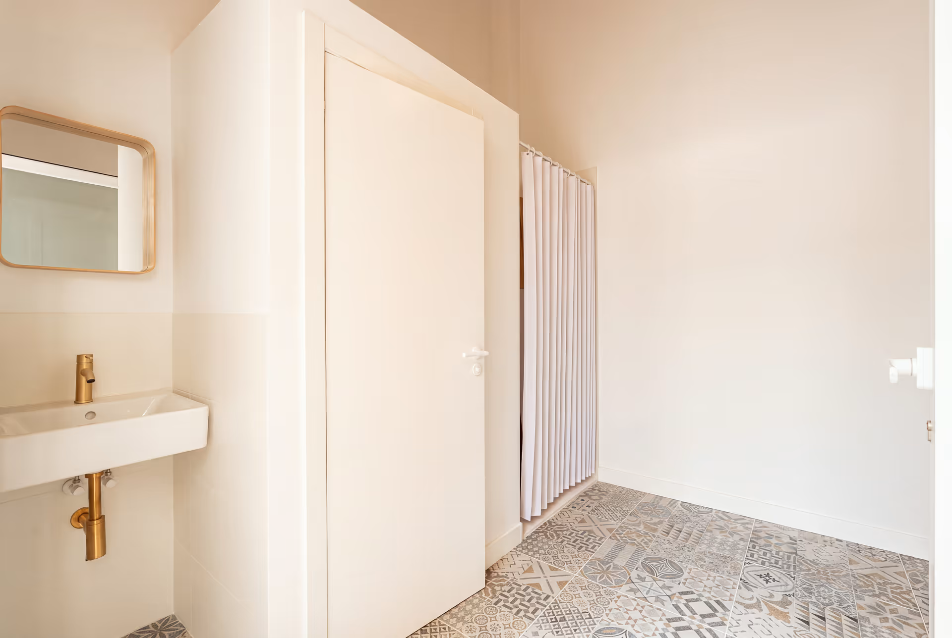 A bright bathroom with patterned floor tiles, a small white sink with brass fittings, and a wood-framed mirror
