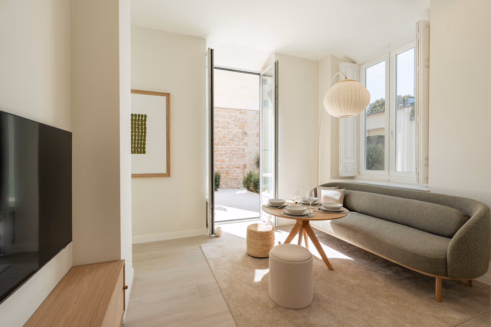 The bright living room of the Oliva residence, with a curved green bouclé sofa and a round wooden dining table. Sunlight streams in through an open door leading to a private stone-walled patio