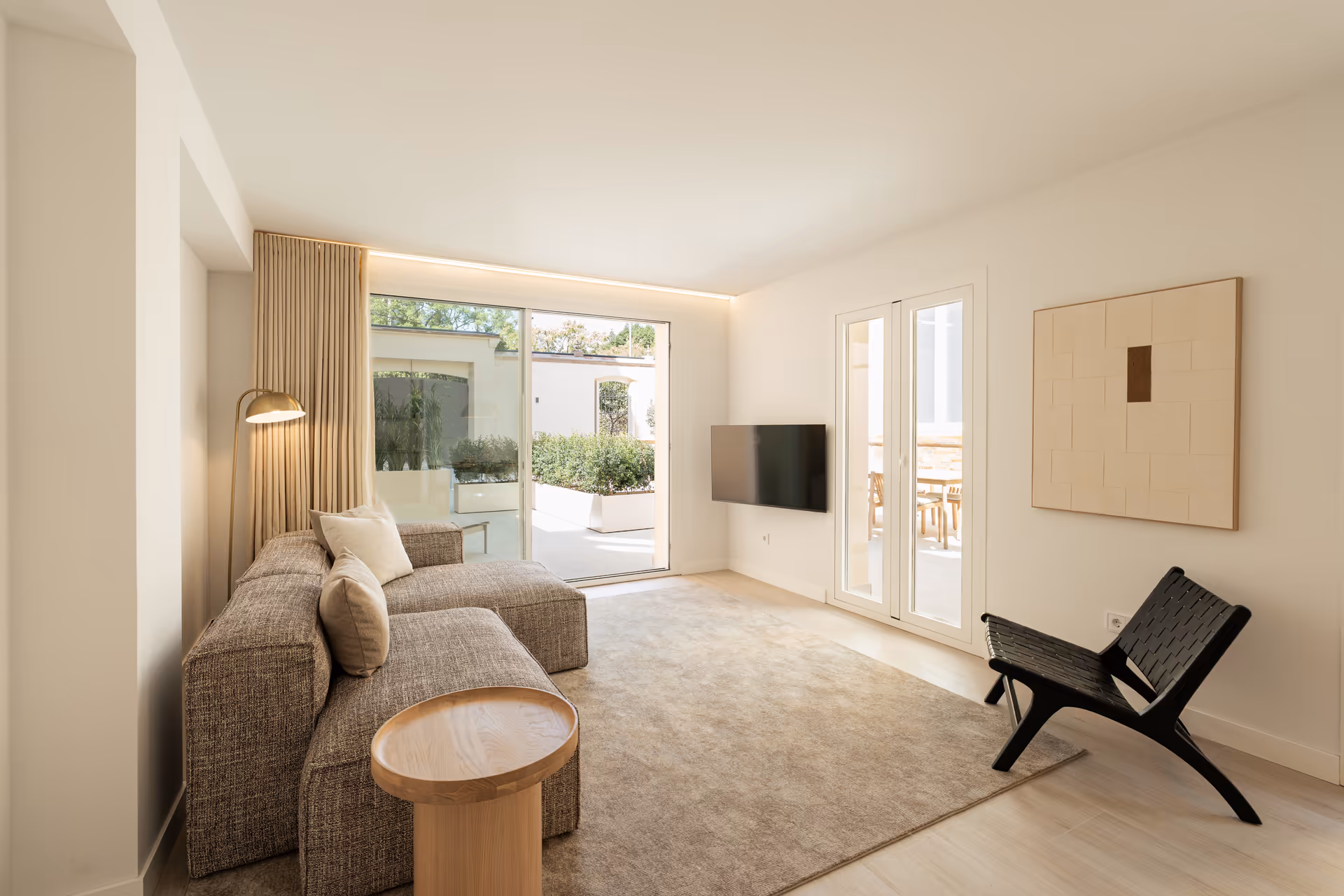 Bright living room in the Toro residence, with a textured L-shaped sofa and a black woven leather chair. Large glass doors open onto a private terrace with planters