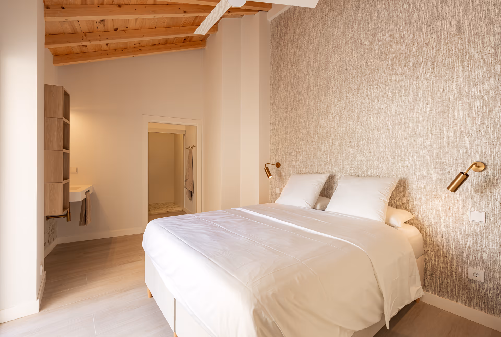 The bedroom in the Toro residence, with a double bed, a sloped wooden ceiling, a textured feature wall and the washbasin and en-suite beyond