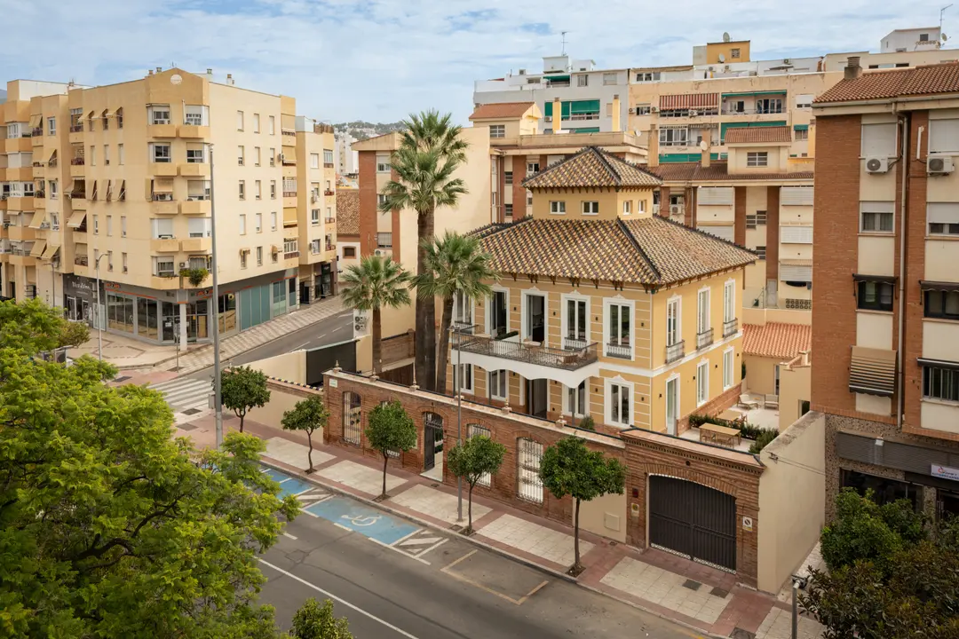 Aerial view of Palacete 10, a restored yellow villa with palm trees, set among modern apartment buildings in Málaga
