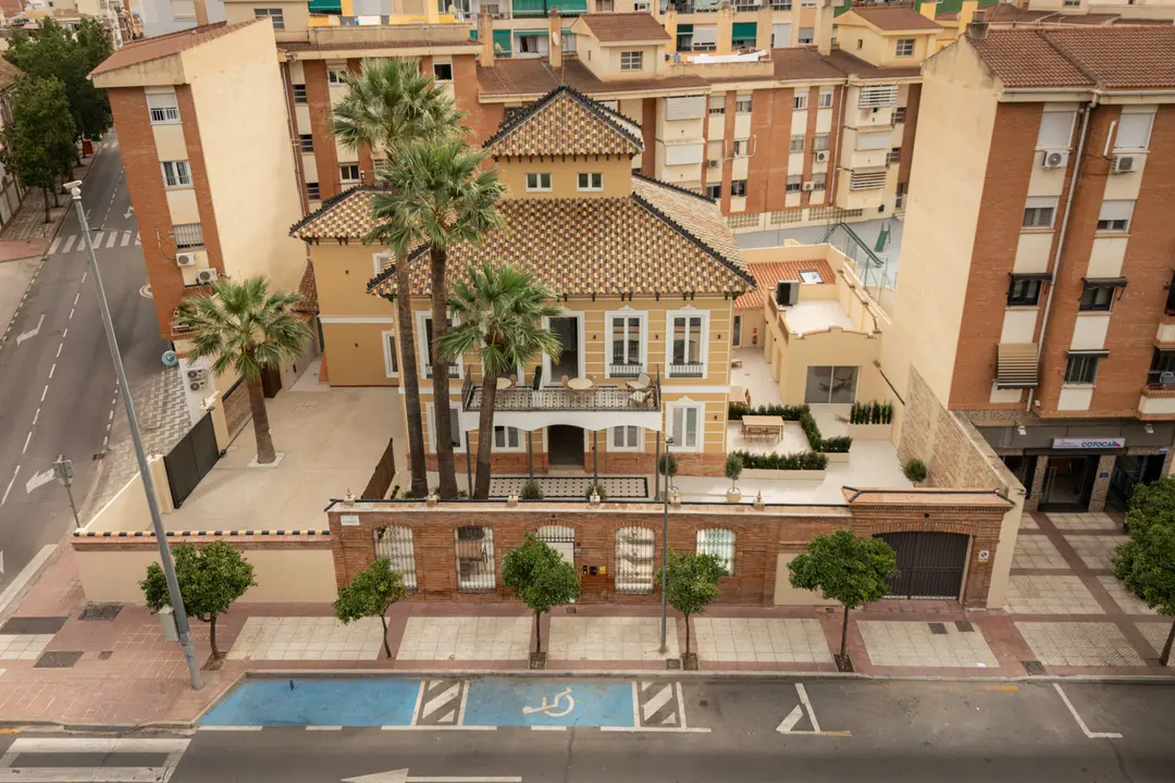 Aerial view of Palacete 10, a restored yellow villa with a tiled roof and palm trees, in its urban Málaga neighbourhood