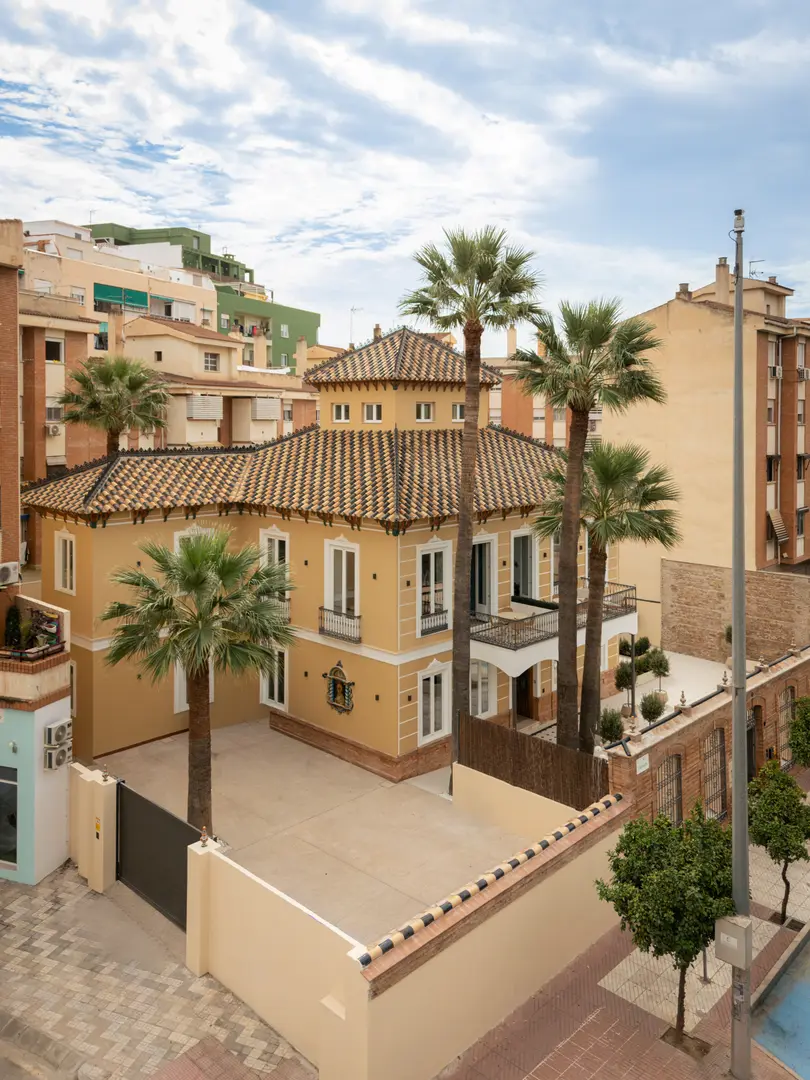 Aerial view of the restored Palacete 10 villa, a yellow building with a tiled roof, surrounded by palm trees in Málaga