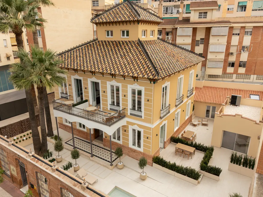 An aerial view of Palacete 10, a restored yellow villa with a tiled roof, tower, and terraces, surrounded by taller buildings in Málaga