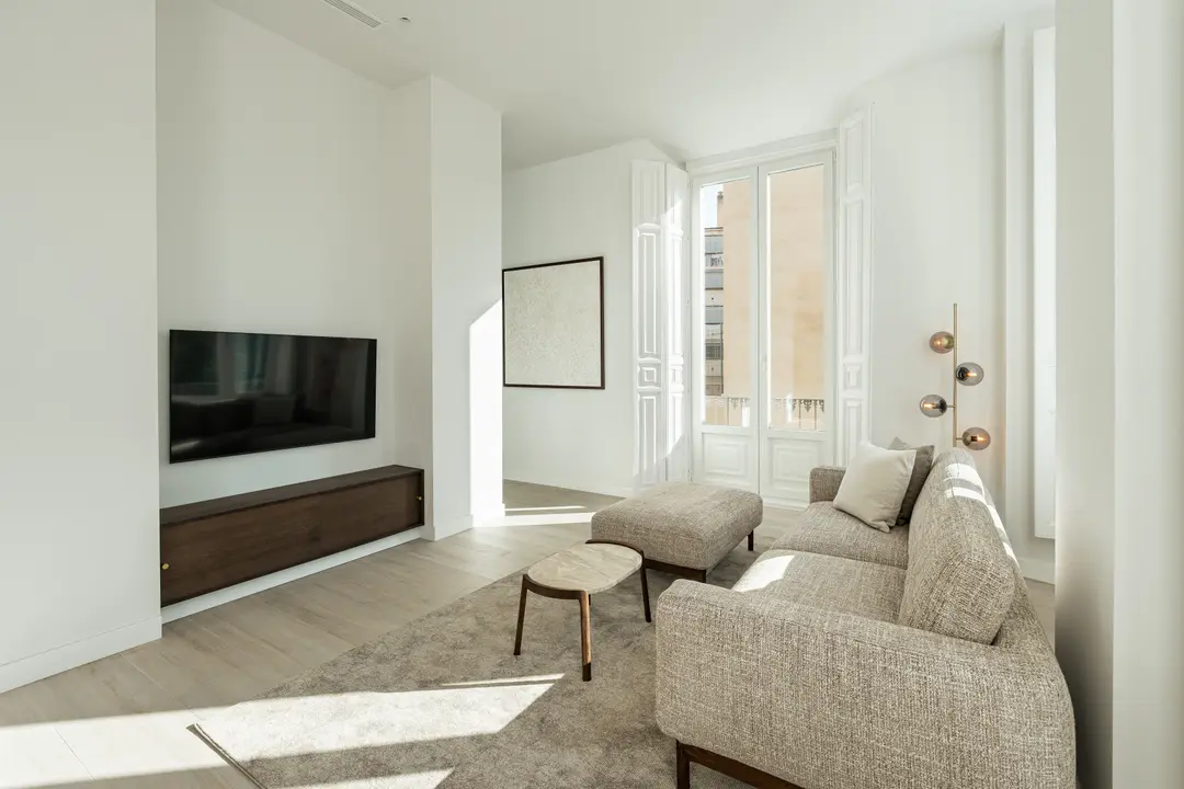 Bright sunlight streams into the living room of the Aurum residence, illuminating a textured beige sofa, light wood floors and a wall-mounted TV.