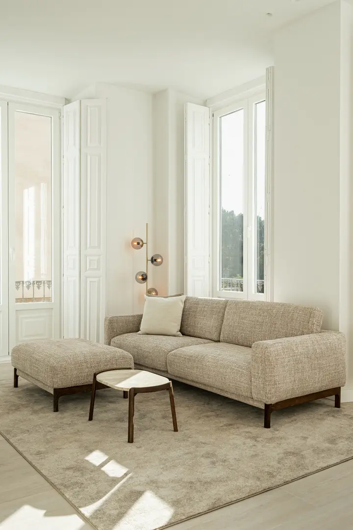 A beige textured sofa and matching footstool in the bright living room of the Aurum residence, next to tall windows with white shutters