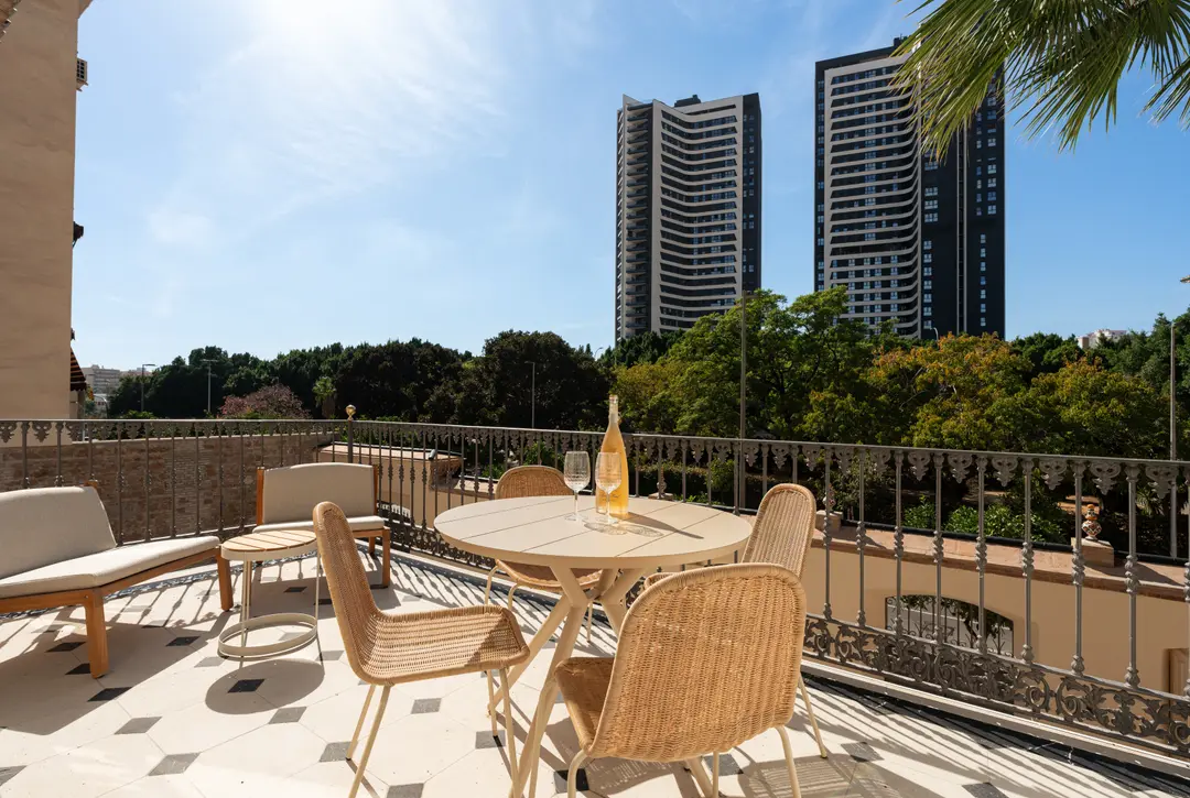 The sunny terrace of the Aurum residence, with a dining table and wicker chairs overlooking trees and modern Málaga towers