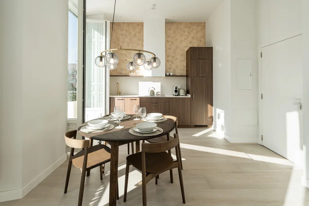 The dining area in the Aurum residence has a dark wood table set for four under a modern chandelier. Sunlight illuminates the open-plan kitchen