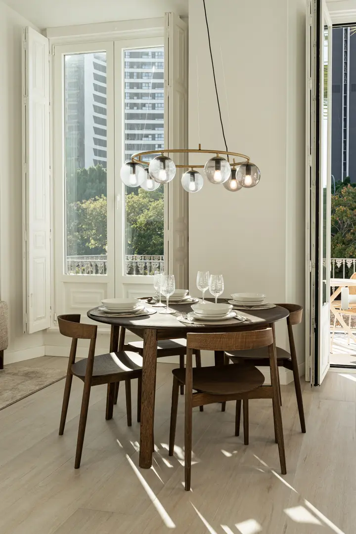 A round dark wood dining table set for four in the Aurum residence, lit by a brass chandelier and bright sunlight from tall windows