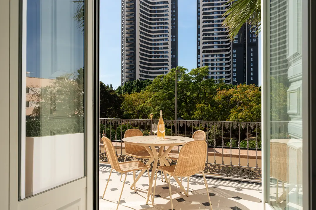 View from inside the Aurum residence onto a sunny terrace with a dining table and chairs overlooking trees and modern towers