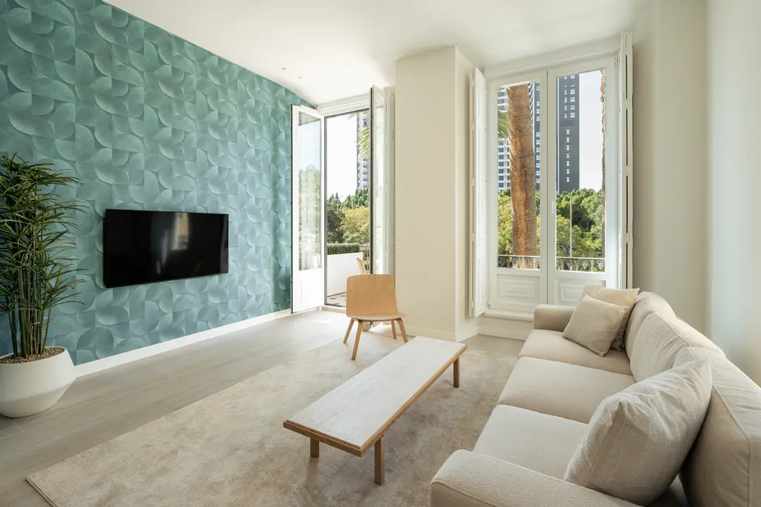 The bright living room of the Azuri residence, featuring a textured teal wall, a beige sofa, and open French doors
