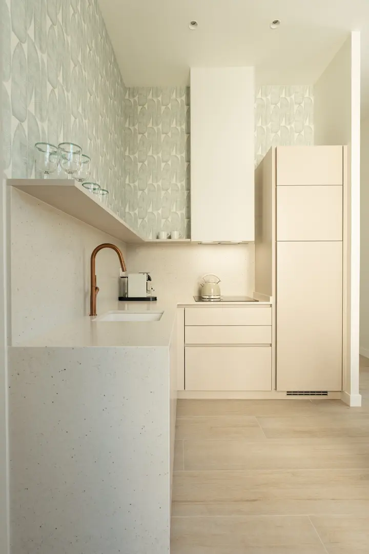 The modern kitchen in the Azuri residence has cream cabinets, a stone worktop, a copper tap, and pale green wallpaper