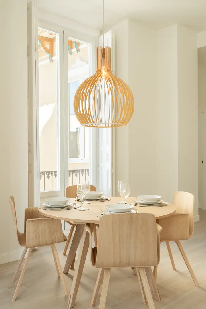 A round wooden dining table set for five in the Azuri residence, lit by a slatted pendant lamp and a tall window