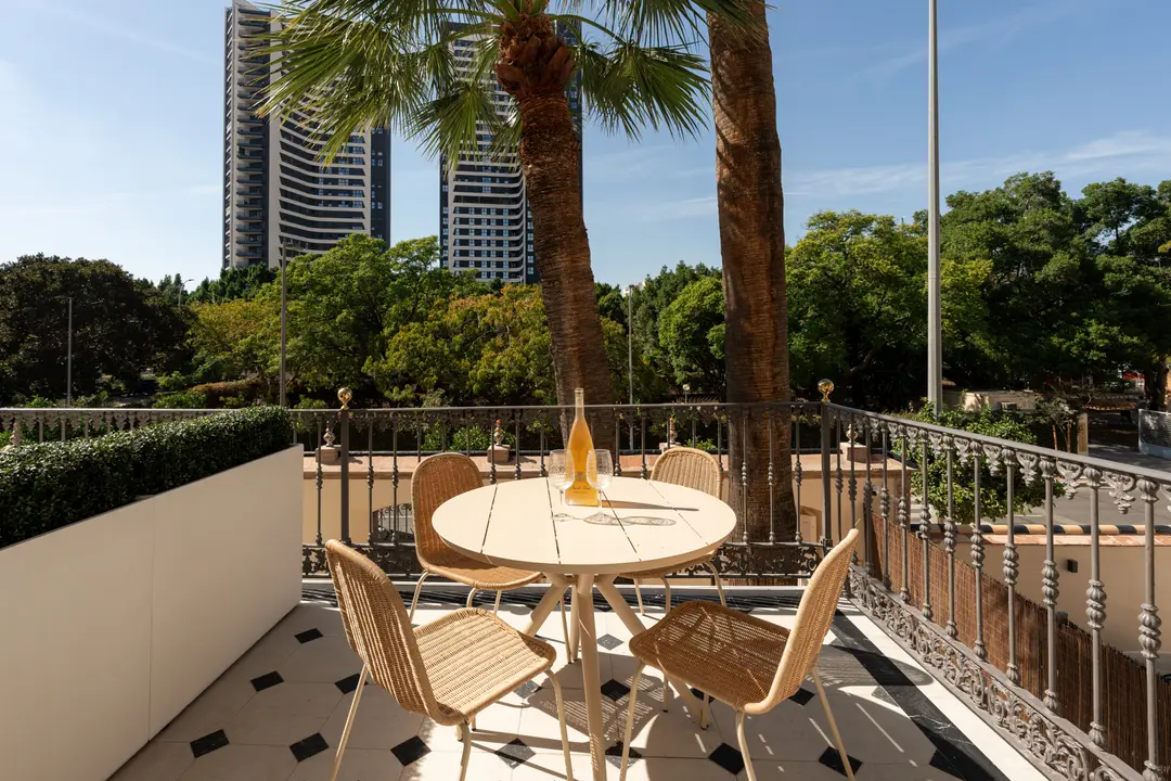 The balcony of the Azuri residence has a round table and four wicker chairs, overlooking palm trees and modern Málaga towers