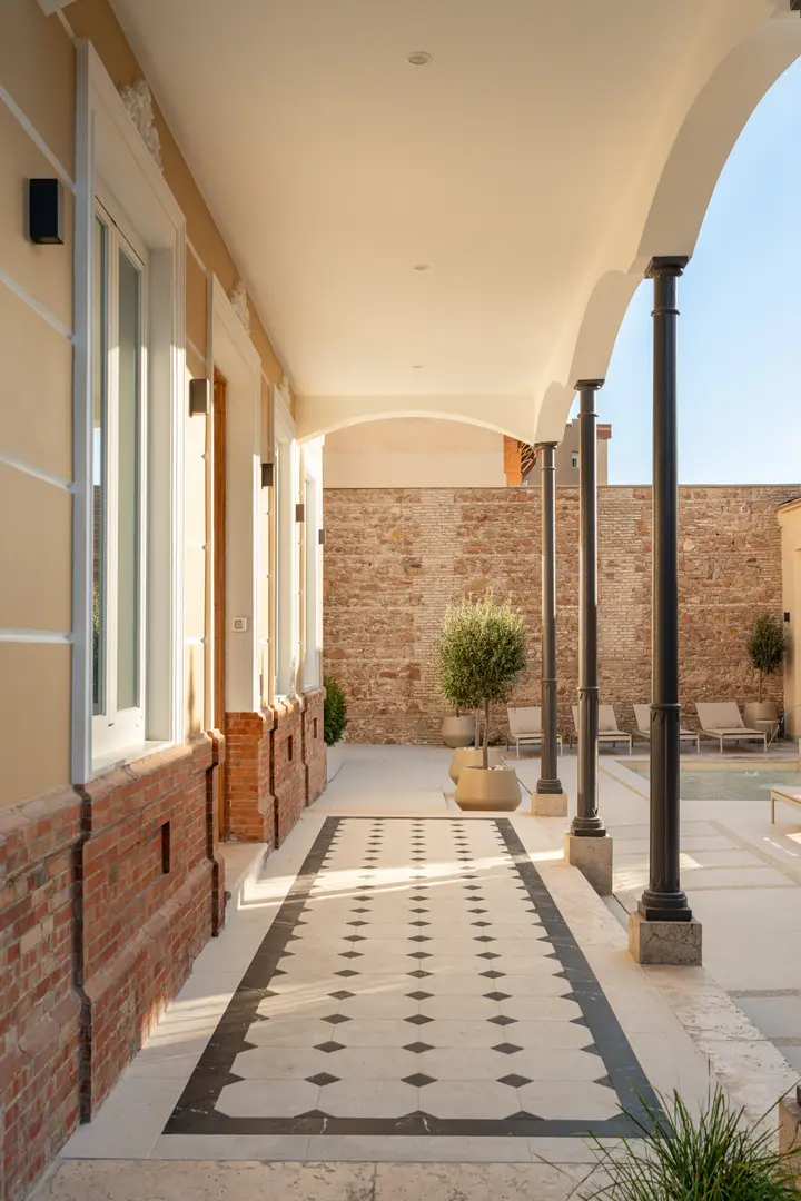 A covered walkway with decorative floor tiles runs alongside the Palacete 10 courtyard with its pond and sun loungers