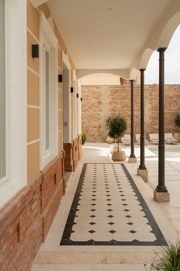 A covered walkway with decorative tiled flooring runs alongside the Palacete 10 courtyard, with its pond and sun loungers