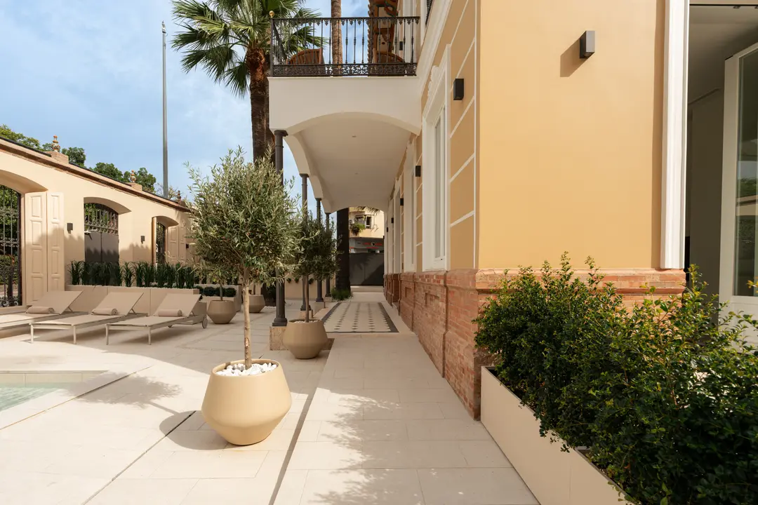 The covered walkway alongside the villa's facade, with potted olive trees lining the path through the courtyard garden towards the pond