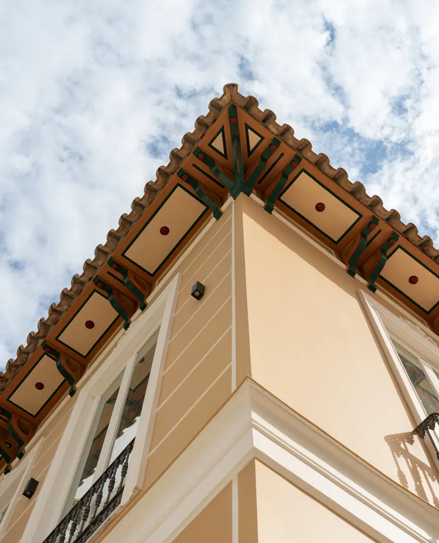 A low-angle view of the decorated wooden eaves and terracotta roof tiles of Palacete 10 against a blue sky