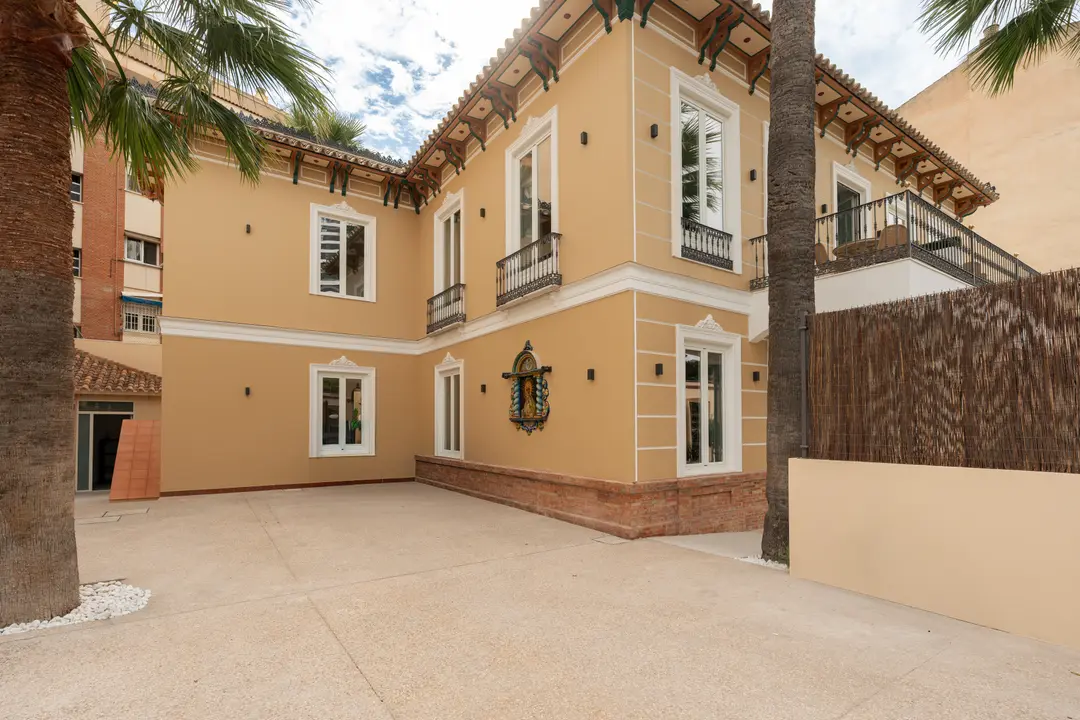 The restored ochre facade of Palacete 10 with its decorative eaves, white windows, and a large paved courtyard with palm trees