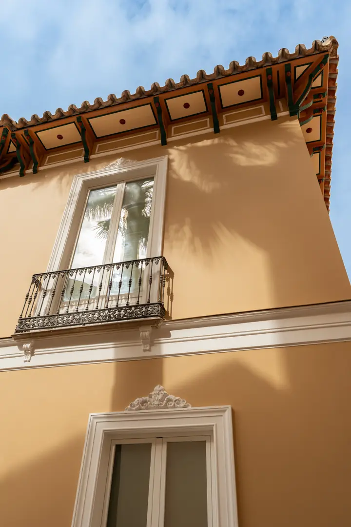 Upward view of the ochre facade of Palacete 10, showing a wrought-iron balcony, decorative eaves, and the shadow of a palm tree against a blue sky