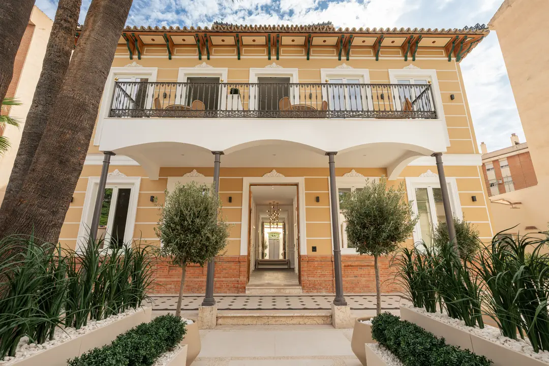 The restored yellow facade of Palacete 10, with its portico, balcony, and landscaped garden with olive and palm trees