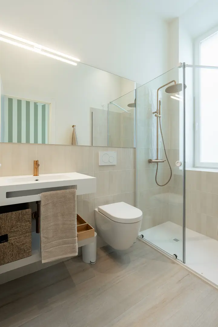 The bathroom in the Oliva residence, with a walk-in shower, wall-hung toilet, and vanity with bronze fittings and wood-effect flooring