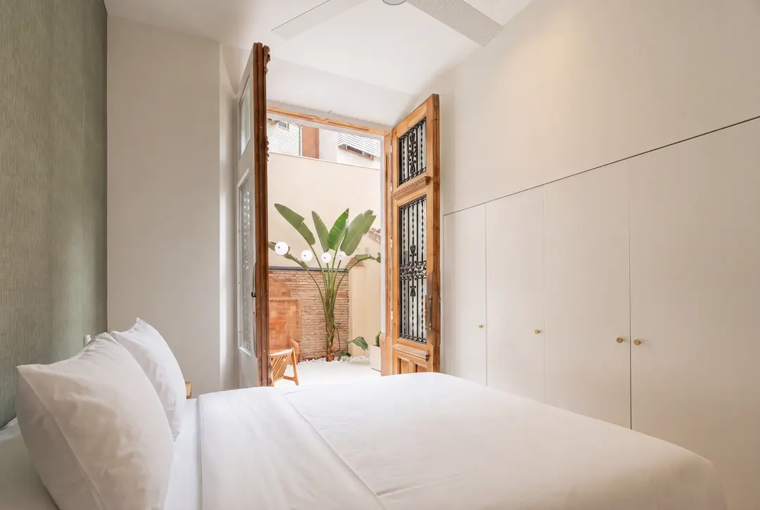 Bedroom in the Oliva residence with a white bed and floor-to-ceiling wardrobes. Tall, antique wooden doors open onto a private terrace