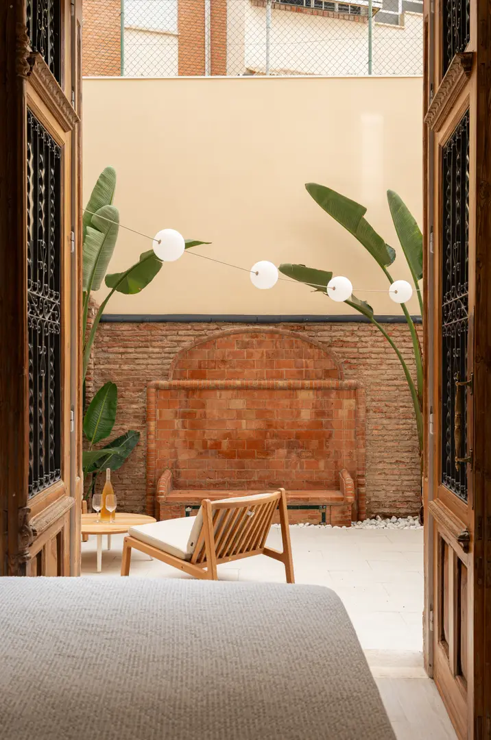 View from the Oliva residence bedroom through open wooden doors onto a private patio with a wooden armchair and terracotta bench