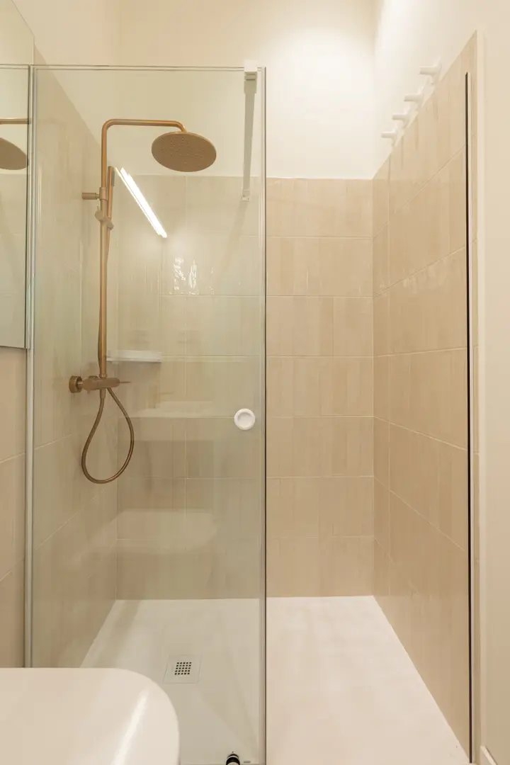 Walk-in shower in the Oliva residence bathroom, featuring sandy-coloured tiles, a glass door, and brushed bronze rain shower fittings