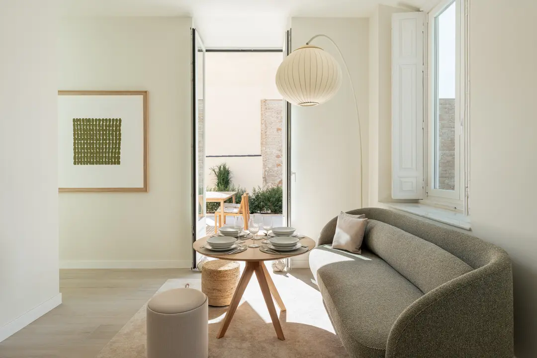 Dining area in the Oliva residence with a curved green sofa and a round wooden table set for four. Doors open onto a sunny private terrace