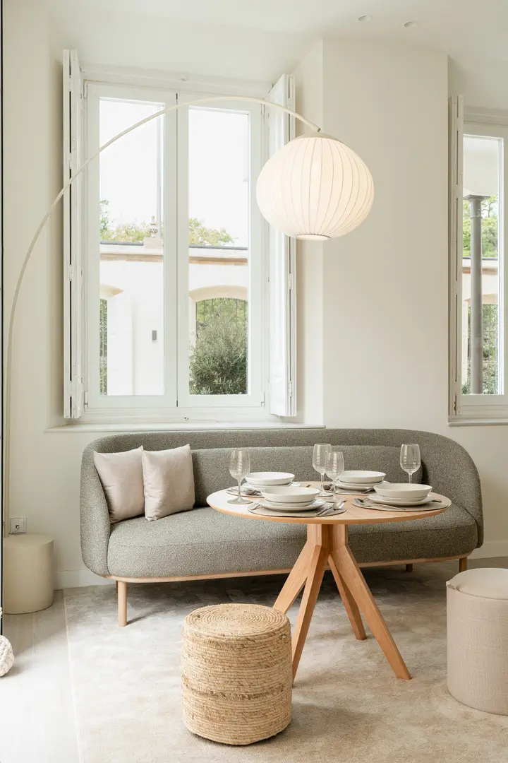 The dining area in the Oliva residence has a round wooden table set for four before a grey sofa, lit by an arc lamp and a tall window