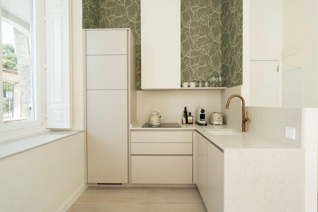 The kitchen in the Oliva residence, with light cabinets, a quartz worktop, a copper tap, and olive-green branch-patterned wallpaper