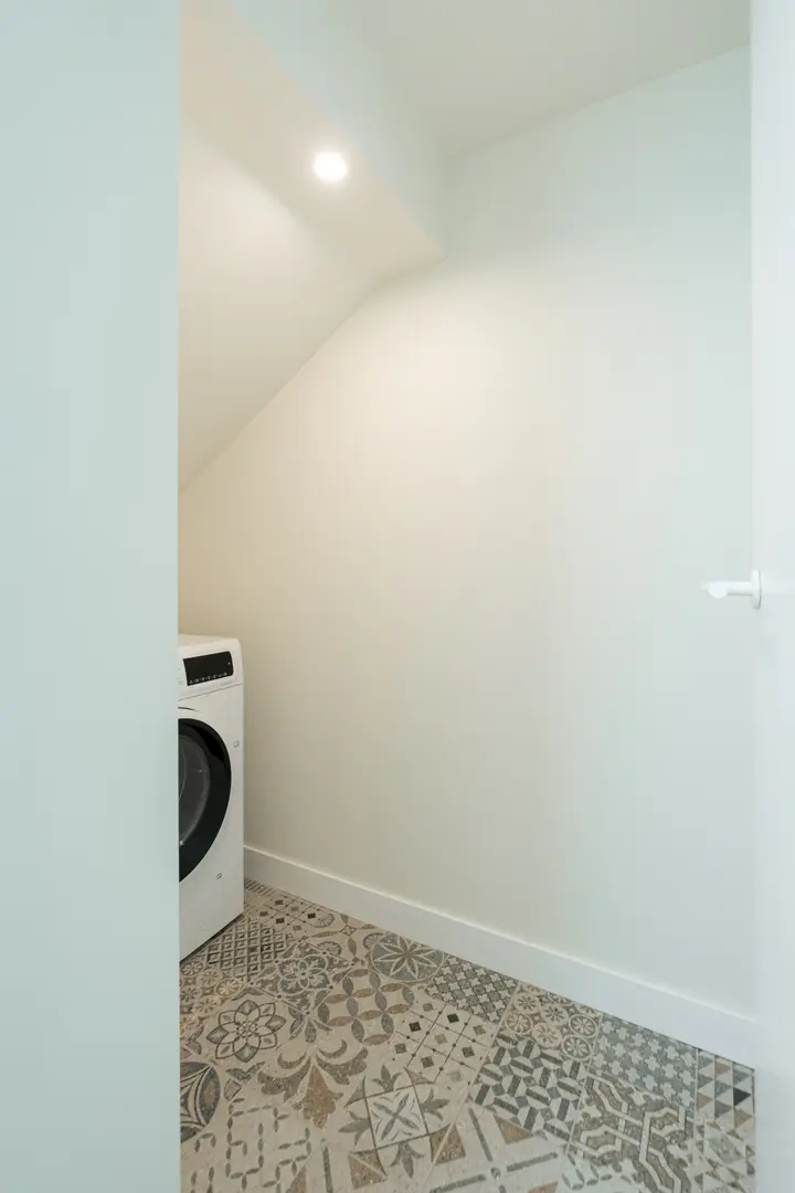 A utility nook in the Oliva residence with a washing machine, sloped ceiling, and a floor of decorative patterned tiles