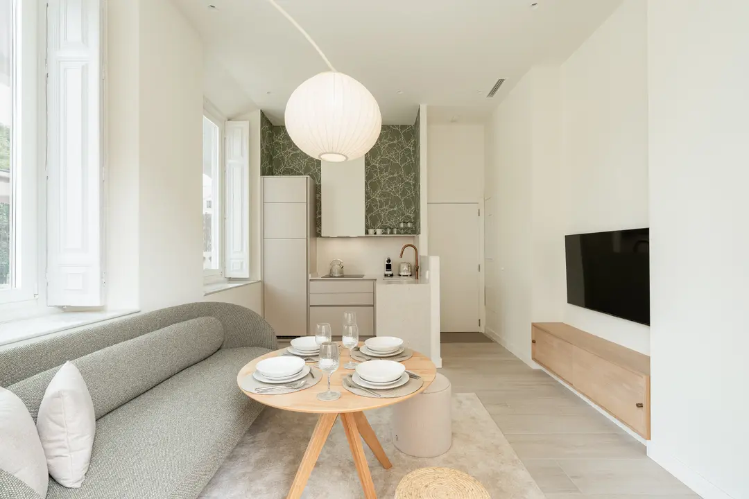 Living area of the Oliva residence with a curved grey sofa, round dining table, and compact kitchen with green wallpaper