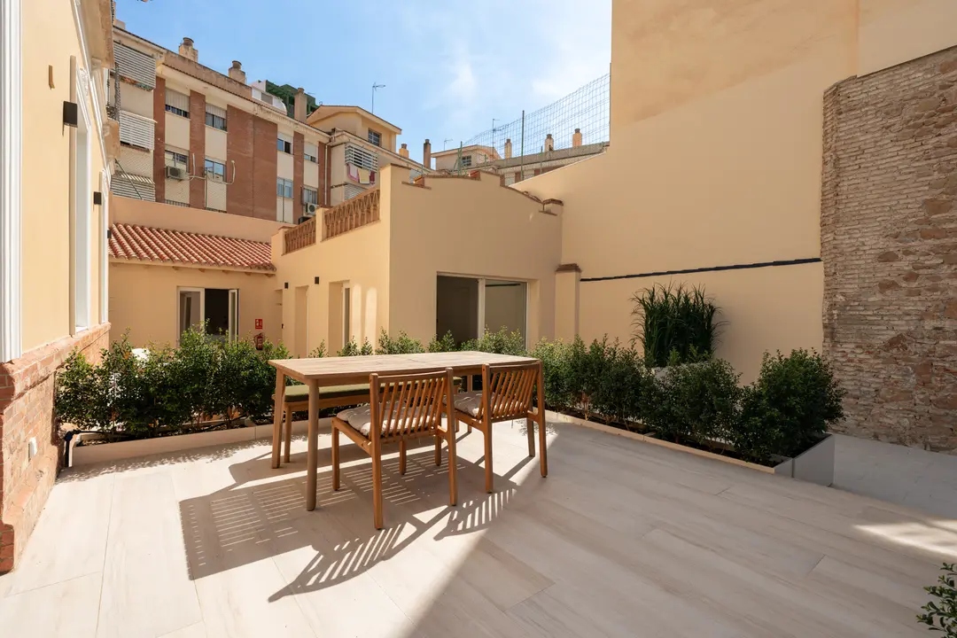 The sunny private terrace of the Oliva residence, with a wooden dining table and chairs on a light tiled floor