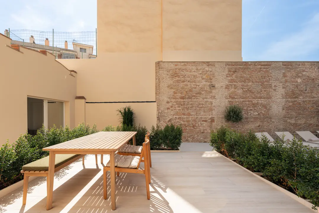 A wooden dining table and bench on the sunny private terrace of the Oliva residence, with green hedges and an exposed brick wall