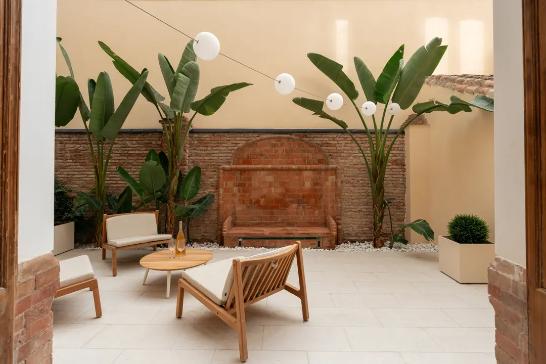 Furnished patio in the Oliva residence with teak armchairs and coffee table, viewed from a doorway, with a terracotta fountain and large plants