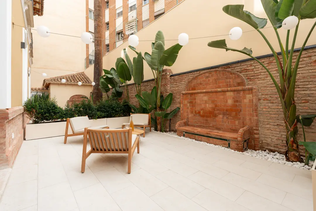 The private patio of the Oliva residence, with wooden armchairs, a coffee table, lush plants, and a decorative brick wall fountain