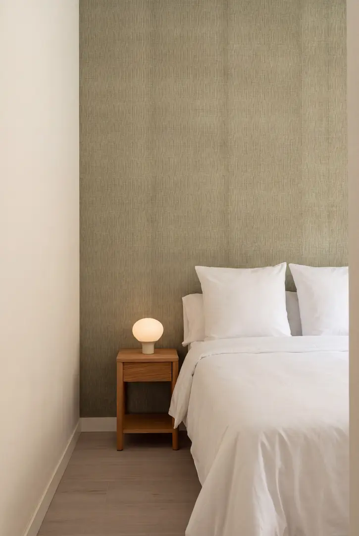 A single bed with a textured white bedspread in the Oliva residence, next to a wooden bedside table with a lamp