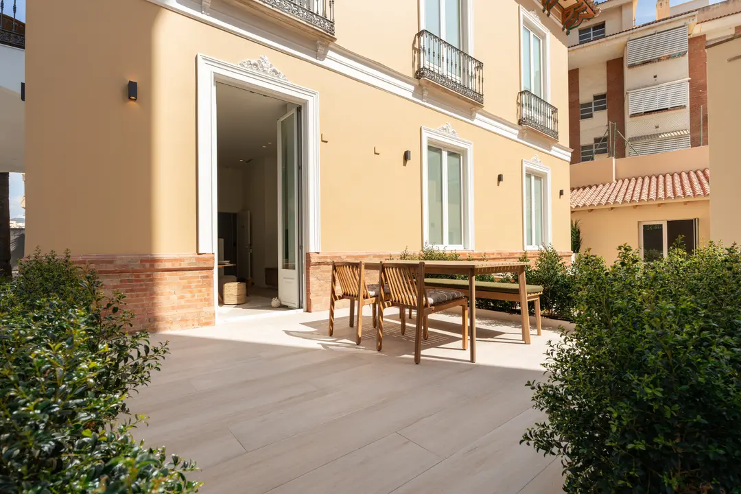 The sunny private terrace of the Oliva residence, with a wooden dining table and chairs on light-coloured paving, surrounded by shrubs