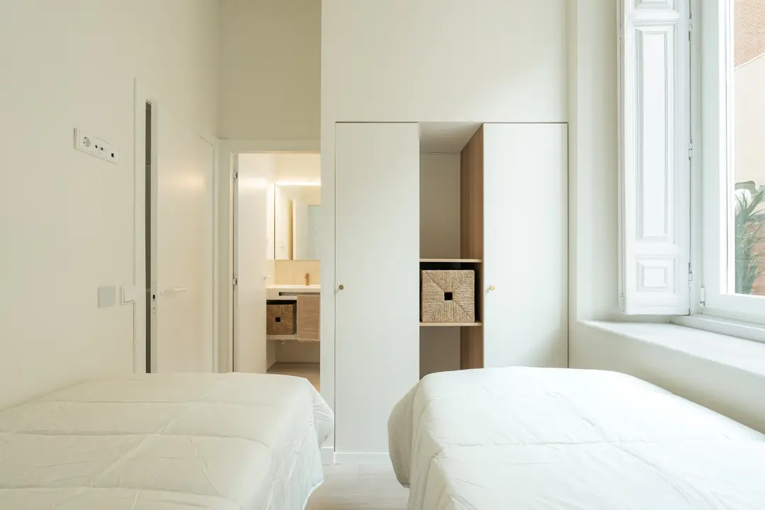 The bright twin bedroom in the Oliva residence has two single beds and a white wardrobe, with a view into the ensuite bathroom