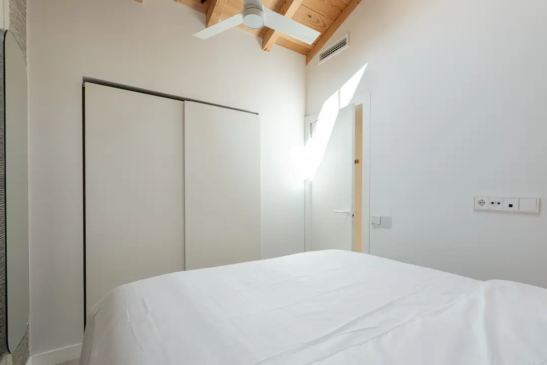 A bright bedroom in the Toro residence with a white bed, a built-in wardrobe and a sloped wooden ceiling with a fan