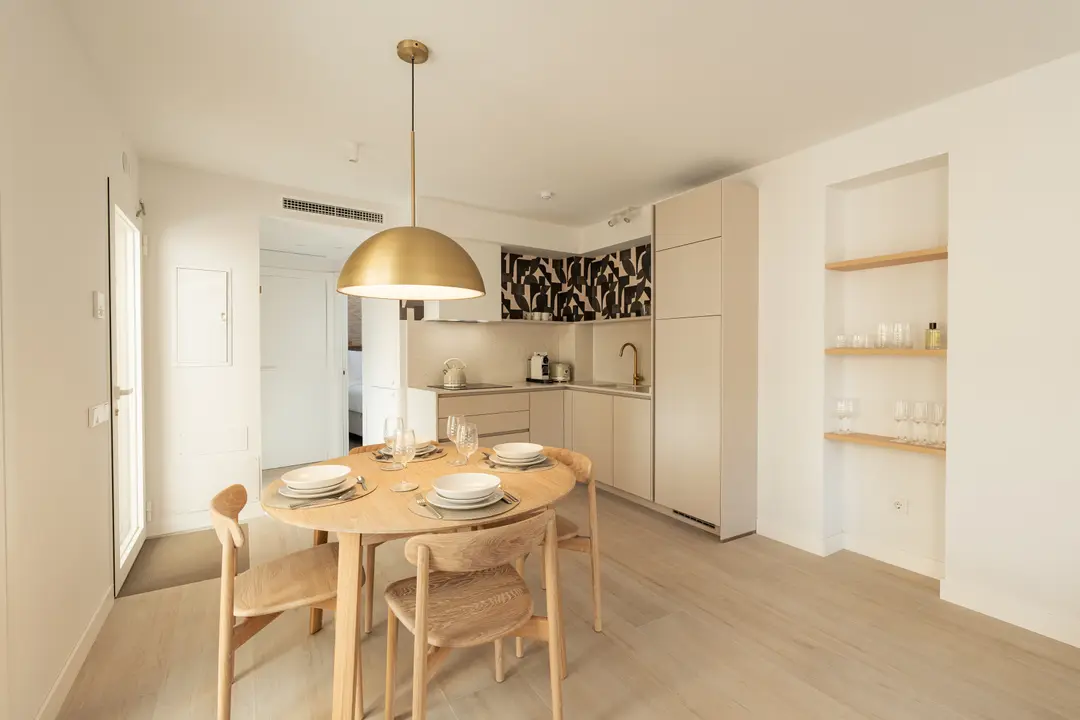 The dining area in the Toro residence, with a round wooden table set for four under a brass pendant lamp. The open-plan kitchen has light cabinets and a patterned backsplash.