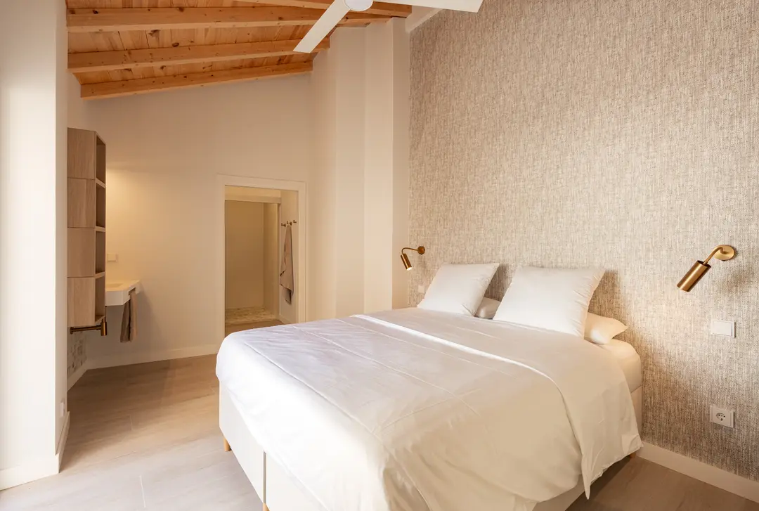 The bedroom in the Toro residence, with a double bed, a sloped wooden ceiling, a textured feature wall and the washbasin and en-suite beyond