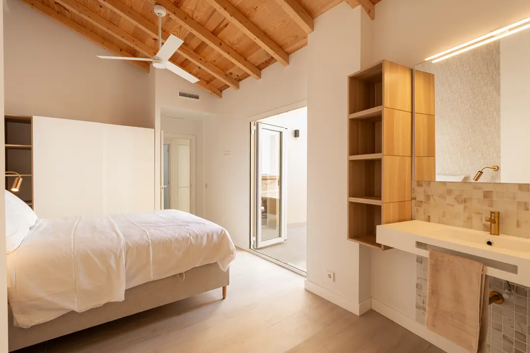 Bedroom in the Toro residence with a pitched wooden ceiling and fan. A double bed faces an integrated sink and a door to a private terrace
