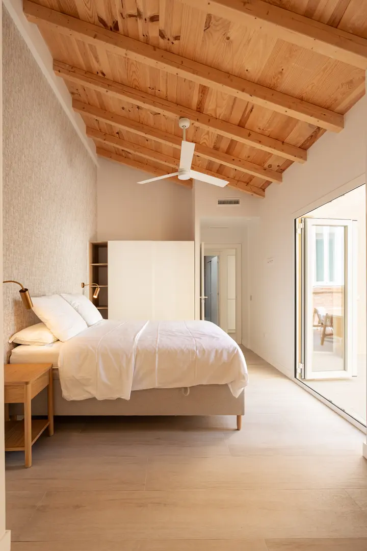 The bright bedroom in the Toro residence has a sloped wooden ceiling, a double bed, and a glass door opening onto a private terrace